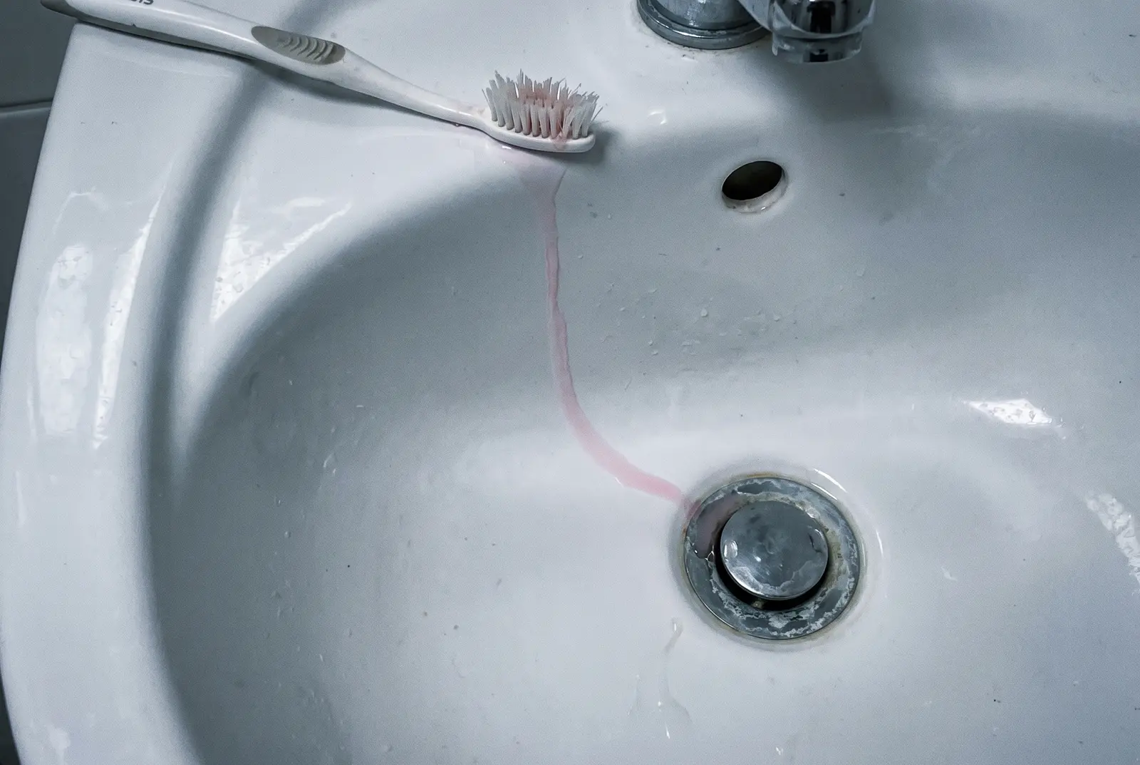 A toothbrush on the edge of a basin, with a thin thread of diluted pink running toward the drain.