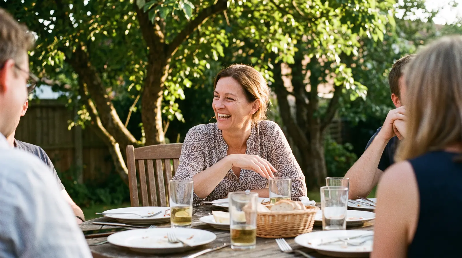 A woman at a backyard table with friends, mid-conversation and clearly amused, afternoon sun through the trees.