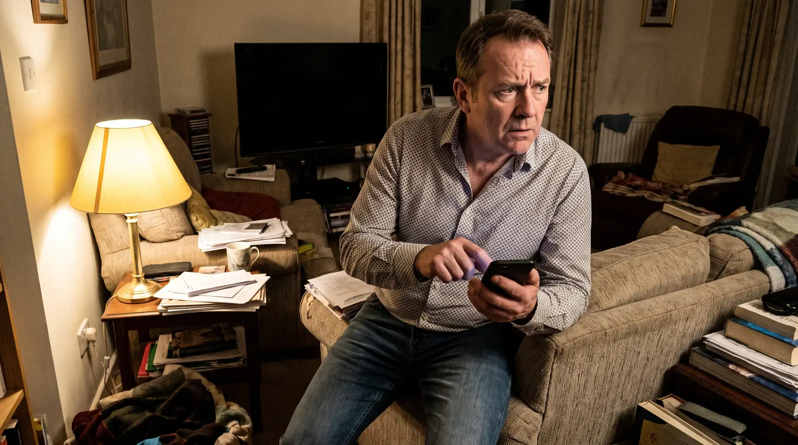 A man sitting alone in his living room at night, phone in both hands, thumb hovering over the screen without pressing.