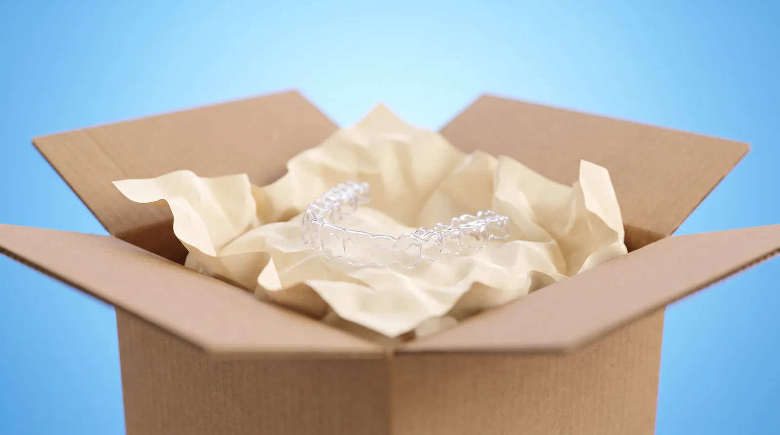 A plain unbranded cardboard box, lid open, with a clear aligner tray loose inside on packing paper