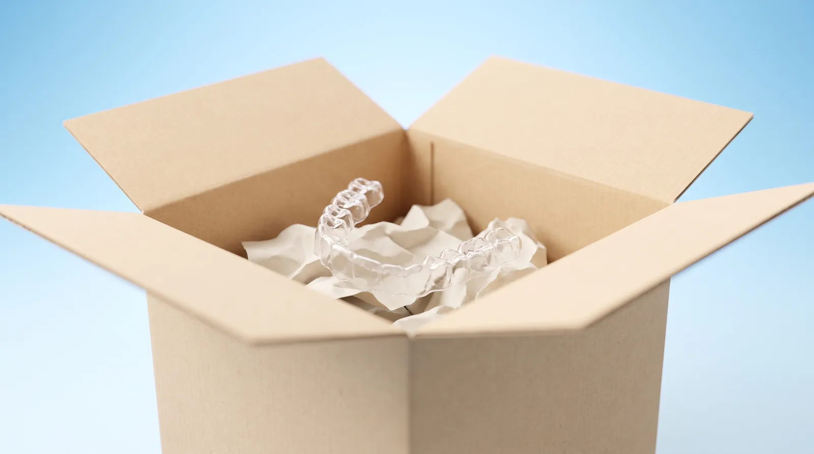 An open cardboard postal box with a clear aligner tray loose inside it